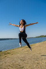 Obraz premium Smiling young female gymnast is jumping in split outdoors near the lake. Healthy lifestyle