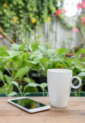 Mobile phone and white coffee cup on wood plank in vegetable farm and flowers garden background