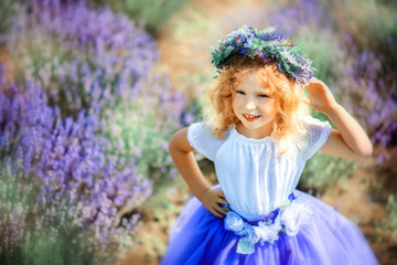 Fototapeta premium Portrait of happy cute little blonde curly girl wearing lavender dress and wreath in lavender field with violet flowers around in sunny summer day. Child in provencal landscape. Close to nature