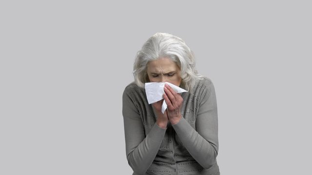 Old senior lady blowing her nose in a napkin. Grey isolated background.