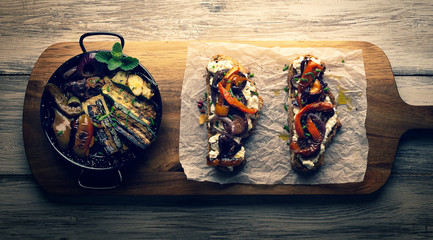Kromki chleba rustykalnego na starej desce z grillowanymi warzywami i ziołami, w stylu vintage i retro, jasne kolory, danie wegetariańskie, widok z góry, płaskie - 334110430