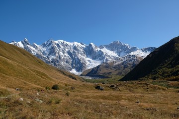 Fototapeta na wymiar Snowy peak Dzhangi-Tau and Shkhara with Khalde Glacier, with green mountainsides and litte wandering tourists on trail in foreground. Greater Caucasus, Svaneti, Georgia