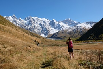 Tourist woman is standing in meadow in valley and taking picture of snowy peaks Dzhangi-Tau and Shkhara with Khalde Glacier. Greater Caucasus, Svaneti, Georgia