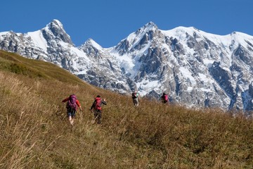 Fototapeta premium Snowy mountains Dzhangi-Tau and Shkhara with Khalde Glacier, with green mountainsides and silhouettes of wandering tourists in foreground. Greater Caucasus, Svaneti, GeorgiaSvaneti
