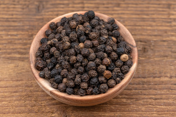 Whole black peppercorns in a small wooden bowl on a rustic wooden background