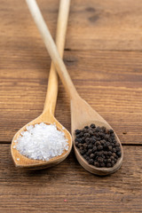 Coarse crystal salt and whole black peppercorns on wooden spoons on a rustic wooden background