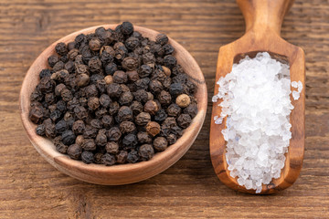 Coarse crystal salt on a small olive wood spice shovel and whole black peppercorns in a small...