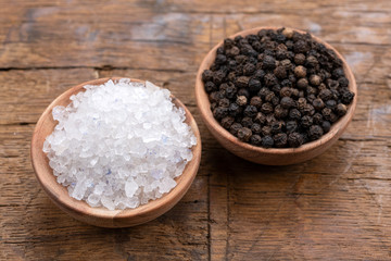 Fototapeta na wymiar Coarse crystal salt and whole black peppercorns in small wooden bowls on a rustic wooden background
