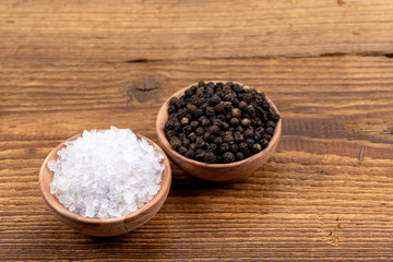 Coarse crystal salt and whole black peppercorns in small wooden bowls on a rustic wooden background