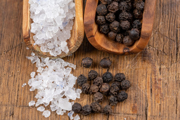 Close-up of coarse crystal salt and whole black peppercorns on small spice shovels made of olive wood on a rustic wooden background