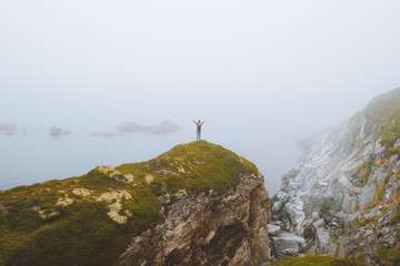 Fototapeta premium Woman standing alone on rocky cliff travel vacation adventure lifestyle outdoor raised hands isolation solitude concept foggy seaside landscape in Norway