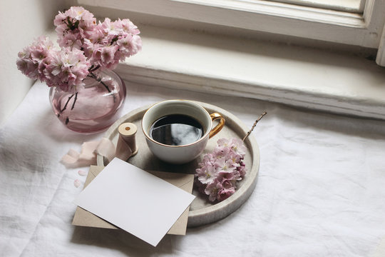 Spring still life scene. Greeting cards mockups, envelope, marble tray, cup of coffee. Vintage feminine styled photo. Floral composition with pink sakura, cherry tree blossoms on table near window.