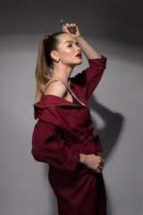 Fototapeta premium A beautiful young woman with red lips and a high tail in a Burgundy raincoat and a black bra. Fashion art Studio portrait