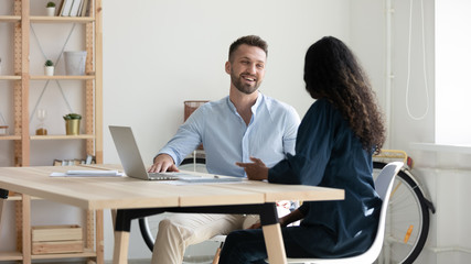 Smiling diverse businesspeople sit at desk brainstorm discuss business ideas together at casual briefing, happy colleagues talk laugh cooperate working on laptop at meeting in office, teamwork concept