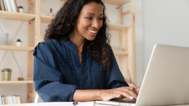 Smiling African American Female Employee Sit At Desk Typing Texting On Modern Laptop In Office, Happy Biracial Woman Worker Busy Working On Computer Gadget, Consult Client Or Customer Online