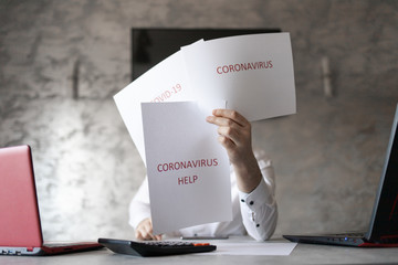 Pracownik w biurze trzymający kartki z napisami coronavirus, covid przed sobą. Biurko z laptopem, dokumetami i kalkulatorem. Coronavirus HELP - obrazy, fototapety, plakaty
