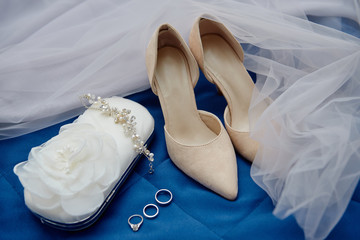 Beige suede female shoes, clutch bag and golden wedding rings on blue background, copy space. Wedding concept