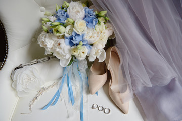 Bridal bouquet of white and blue flowers with satin ribbon, beige women shoes, two golden wedding rings and purple dress on armchair, copy space. Wedding concept