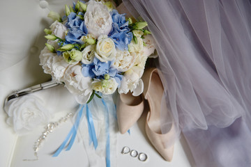 Bridal bouquet of white and blue flowers with satin ribbon, beige women shoes, two golden wedding rings and purple dress on armchair, copy space. Wedding concept