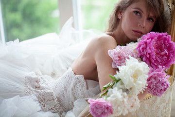 Bride in beautiful dress lying on sofa whit peonies in white studio interior. Romantic wedding style shot with sun light