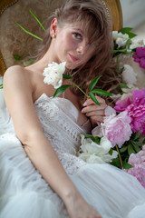 Bride in beautiful dress lying on sofa whit peonies in white studio interior. Romantic wedding style shot with sun light