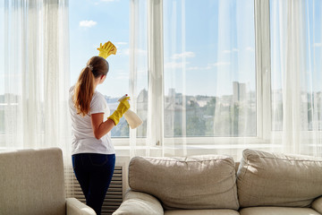 Woman in white shirt and yellow rubber gloves cleaning window with cleanser sprayer and yellow rag...