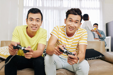 Excited Vietnamese guys playing video games at home, their friends are talking in background