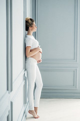 A pregnant European girl stands by the window. Profile shot. Studio photo shoot. Soft focus.