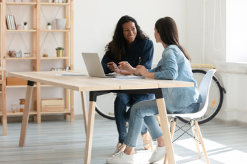 Smiling female colleagues sit at desk in office talk brainstorm work together on laptop, happy woman coworkers businesspeople speak discuss business ideas collaborate on modern computer at meeting