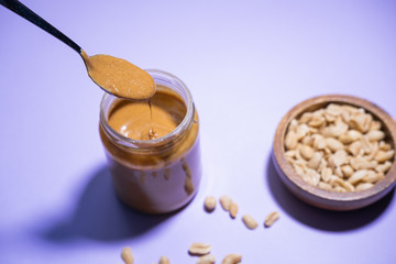 Spoon full of peanut butter, falling into a glass jar on purple background. Vegan product.