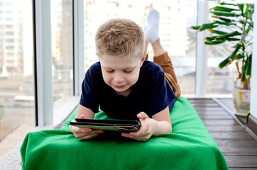 Child boy reads an e-book on a tablet and watches cartoons. Isolation from the outside world. World Quarantine, Coronovirus Pantemia, COVID-19. Close-up. The child is sitting on the balcony. To stay