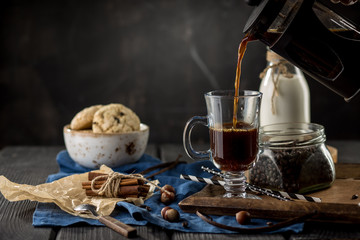 pouring coffee in cup with coffee beans, spoon, cinnamon sticks, bottle of milk and cookies on black background