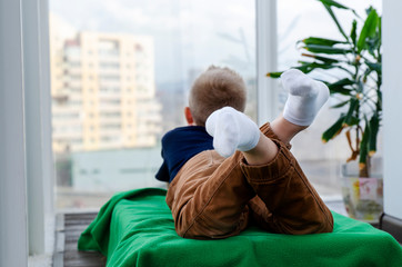 Child boy lies on a blanket on the balcony and looks out the window. Isolation from the outside world. World Quarantine, Coronovirus Pantemia, COVID-19. To stay home. Preventive measures. Danger