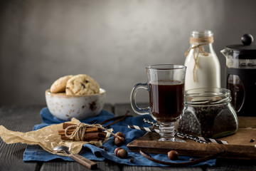 Cup of coffee with coffee beans, spoon, cinnamon sticks, bottle of milk and cookies on dark background