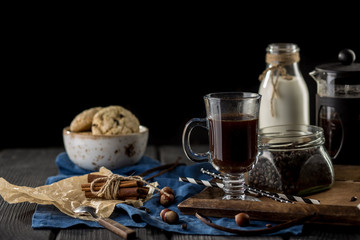 Cup of coffee with coffee beans, spoon, cinnamon sticks, bottle of milk and cookies on black background