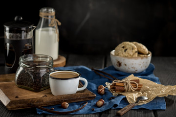 Cup of coffee with coffee beans, spoon, cinnamon sticks, bottle of milk and cookies on black background