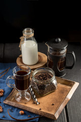 Cup of coffee with coffee beans, spoon, cinnamon sticks, bottle of milk and cookies on black background