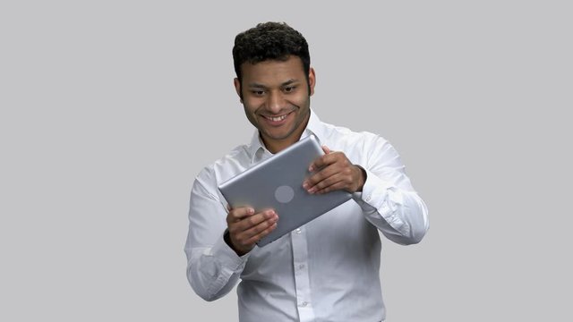 Very exctied dark-skinned guy playing video game on his tablet pc. Infantile man's killing time. Grey isolated background.