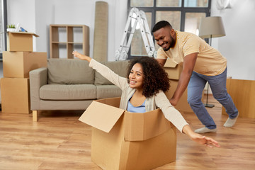 Fototapeta na wymiar moving, repair and real estate concept - happy african american couple having fun and playing with cardboard box at new home