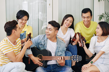 Positive friends clinking bottles of beer when their friend playing guitar and singing for them