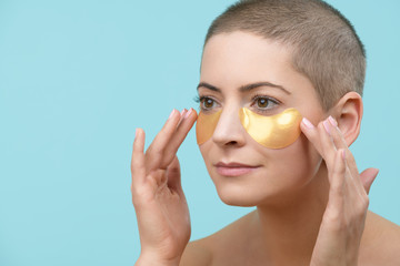 Attractive young woman applying gold collagen eye mask. Photo of mid adult caucasian woman with shaved head and healthy skin isolated on pastel blue background. Beauty products concept.