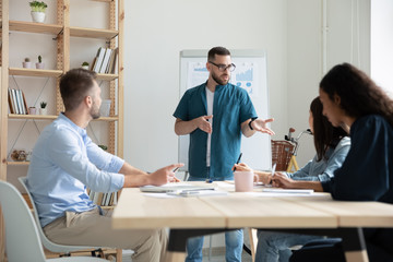 Concentrated Caucasian male leader in glasses stand lead meeting with diverse colleagues, focused confident young boss head briefing with coworkers in office, cooperation, teamwork concept