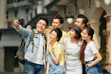 Happy Vietnamese young man taking selfie with group of friends when standing in the street