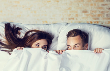 Picture of attractive couple peeping from bedsheet on the bed at bedroom. Love, relationship, dating, happy people, bedtime concept.