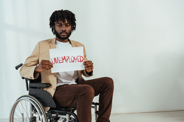 Disabled african american man looking at camera and holding placard with unemployed lettering on wheelchair on white