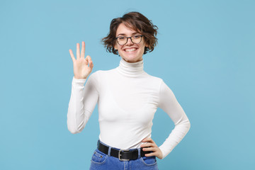 Fototapeta premium Smiling young brunette woman girl in casual white clothes, eyeglasses posing isolated on pastel blue wall background studio portrait. People lifestyle concept. Mock up copy space. Showing OK gesture.
