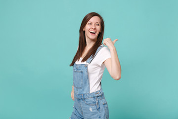 Side view of laughing young brunette woman girl in casual denim clothes posing isolated on blue turquoise wall background in studio. People lifestyle concept. Mock up copy space. Pointing thumb aside.