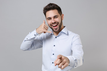 Cheerful young unshaven business man in light shirt isolated on grey background. Achievement career wealth business concept. Doing phone gesture like says call me back pointing index finger on camera.