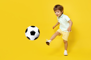 Charming little kid in casual summer outfit play with soccer ball over yellow background.