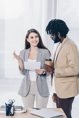 Fototapeta premium Recruiter with pen and paper looking at african american employee with disposable cup of coffee and smiling near table